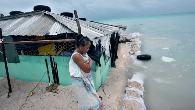 Eine Frau steht auf dem Festland und schaut mit verschränkten Armen auf die Wassermassen des Meeres, die bereits ihr Haus erfasst haben. Eine Frau steht auf dem Festland und schaut mit verschränkten Armen auf die Wassermassen des Meeres, die bereits ihr Haus erfasst haben.