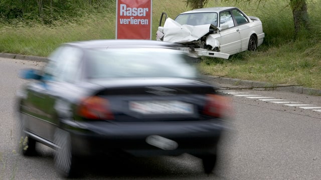 Auto fährt auf einer Strasse. Dahinter ist ein Schild mit der Aufschrift «Raer verlieren» und ein beschädigtes Auto zu sehen.