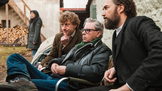 Drei Männer unterschiedlichen Alters sitzen an einem Filmset und blicken konzentriert drein. Drei Männer unterschiedlichen Alters sitzen an einem Filmset und blicken konzentriert drein.