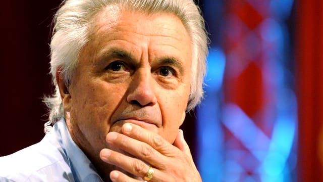 Ein Mann mit weissem Haar schaut nachdenklich, die eine Hand ans Kinn gelegt. Ein Mann mit weissem Haar schaut nachdenklich, die eine Hand ans Kinn gelegt.