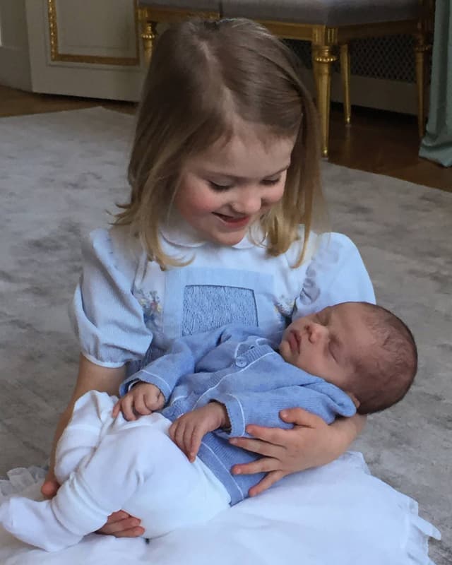 Prinzessin Estelle sitz in einem weiss-blauen Kleidchen am Boden und hält ihren Baby-bruder Oscar im Arm. Prinzessin Estelle sitz in einem weiss-blauen Kleidchen am Boden und hält ihren Baby-bruder Oscar im Arm.