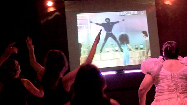 Ein Video auf der Leinwand zeigt einen Mann mit ausgebreiteten Armen. Die Menschen davor machen die selbe Geste. Ein Video auf der Leinwand zeigt einen Mann mit ausgebreiteten Armen. Die Menschen davor machen die selbe Geste.