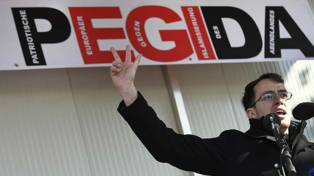 Ignaz Bearth davos in microfon ed avant in transparent pendi si sur el cun scret si PEGIDA. El tegna ses maun dretg en l'aria sco per engirar. Ignaz Bearth davos in microfon ed avant in transparent pendi si sur el cun scret si PEGIDA. El tegna ses maun dretg en l'aria sco per engirar.