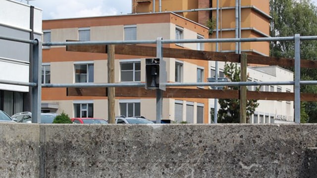 Brücke mit Metallgeländer, daran ist ein Messgerät befestigt. Dahinter sind Häuser zu sehen. Brücke mit Metallgeländer, daran ist ein Messgerät befestigt. Dahinter sind Häuser zu sehen.