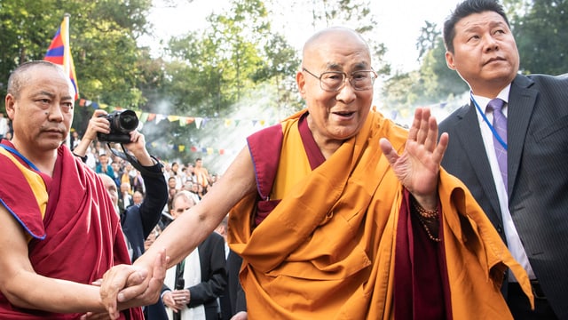 Der Dalai Lama in orangefarbenem Gewand wird begleitet von zwei Personen und winkt Menschen zu. Der Dalai Lama in orangefarbenem Gewand wird begleitet von zwei Personen und winkt Menschen zu.