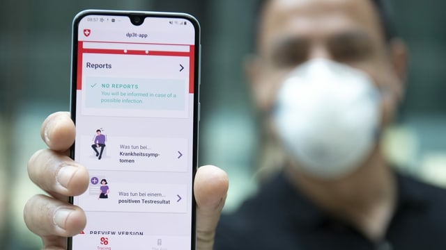Ein Mann mit Gesichtsmaske hält ein Smartphone in die Kamera, auf der eine App läuft. Ein Mann mit Gesichtsmaske hält ein Smartphone in die Kamera, auf der eine App läuft.