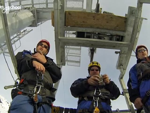 Drei Seilbahn-Mechatroniker auf einer Arbeitsplatform unter einem Seilbahnmast.