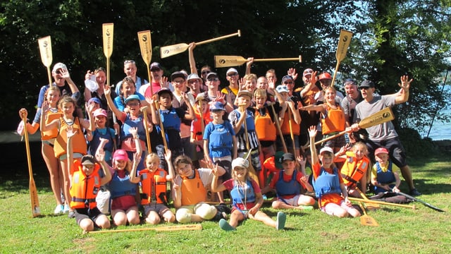 Eine 30-köpfige Gruppe steht auf einer Wiese, alle tragen eine orange Schwimmweste und heben mit den Händen Ruder in die Höhe. Eine 30-köpfige Gruppe steht auf einer Wiese, alle tragen eine orange Schwimmweste und heben mit den Händen Ruder in die Höhe.