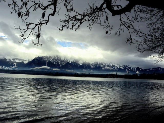 Im Vordergrund ist der Thunersee zu sehen. Die Landschaft ist wolkenverhangen. Einzig in der Distanz ist eine kleine blaue Lücke in den Wolken zu sehen.