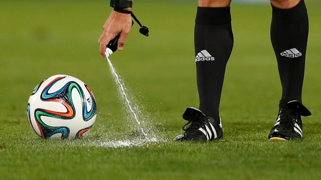 Schiedsrichter sprayt Schaum auf dne Rasen, daneben ein Fussball. Schiedsrichter sprayt Schaum auf dne Rasen, daneben ein Fussball.