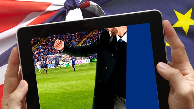 Auf einem Tablet ist ein Fussballtrainer am Spielfeldrand zu sehen. Hinter dem Tablet sind eine europäische und eine UK-Fahne zu sehen.