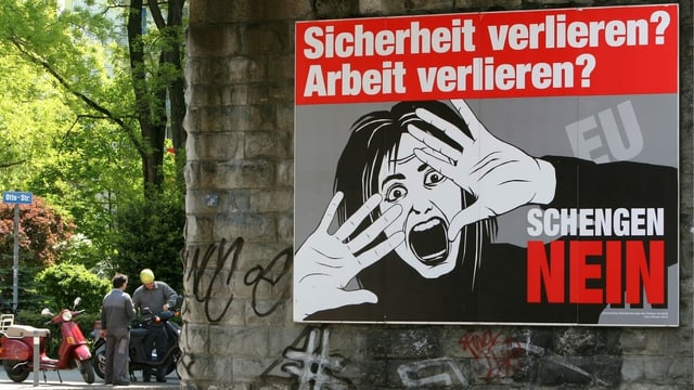 Zwei Männer begutachten eine Vespa hinter der Kulisse eines riesigen Schengen-Nein Plakates. Zwei Männer begutachten eine Vespa hinter der Kulisse eines riesigen Schengen-Nein Plakates.