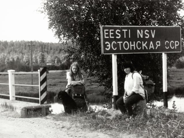 Altes Foto: Zwei Frauen sitzen vor einer Brücke mit Strassenschild. Altes Foto: Zwei Frauen sitzen vor einer Brücke mit Strassenschild.