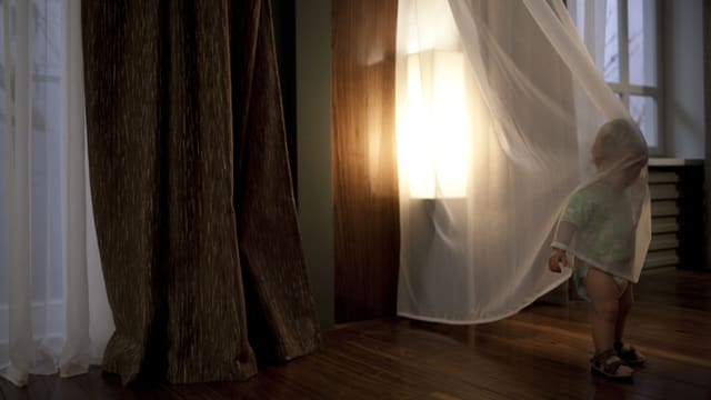 Ein kleines Kind umhüllt sich mit einem durchsichtigen Vorhang, wobei der Kopf und der Oberkörper bedeckt sind. Hinter dem Kind scheint die Sonne durch ein grosses Fenster. Ein kleines Kind umhüllt sich mit einem durchsichtigen Vorhang, wobei der Kopf und der Oberkörper bedeckt sind. Hinter dem Kind scheint die Sonne durch ein grosses Fenster.
