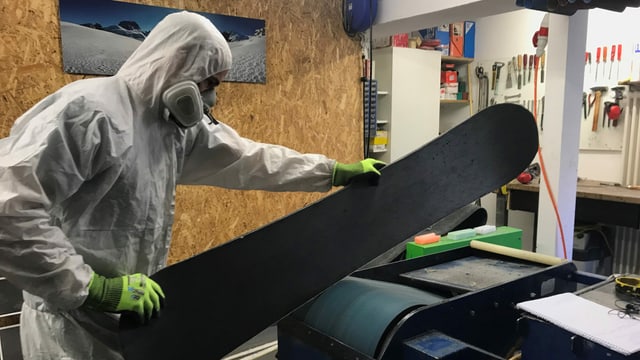 Ein Mann in weissem Overall und mit Atemschutz hält ein Snowboard auf eine Schleifmaschine Ein Mann in weissem Overall und mit Atemschutz hält ein Snowboard auf eine Schleifmaschine