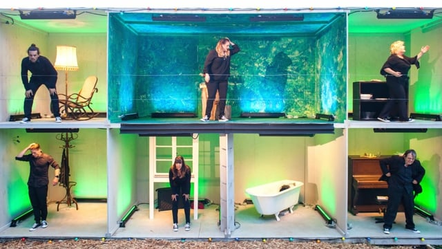 Sechs Schauspieler auf einer Bühne, die einem Puppenhaus ähnelt. Sechs Schauspieler auf einer Bühne, die einem Puppenhaus ähnelt.