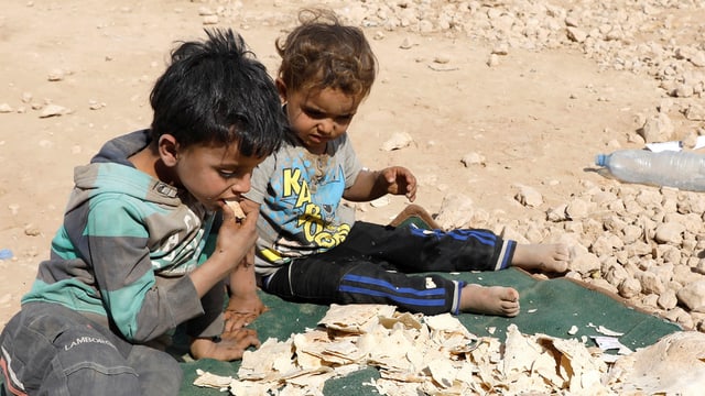 Zwei Kinder sitzen auf steinigem Boden auf einer grünen Decke, auf der Brot trocknet. Eines der Kindert kostet ein Brotstück. Zwei Kinder sitzen auf steinigem Boden auf einer grünen Decke, auf der Brot trocknet. Eines der Kindert kostet ein Brotstück.