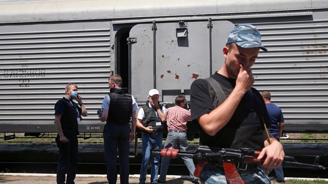 Mann mit Gewehr hält sich die Nase zu. Dahinter ist eine Gruppe Personen vor einem Eisenbahnkühlwagen zu sehen. Mann mit Gewehr hält sich die Nase zu. Dahinter ist eine Gruppe Personen vor einem Eisenbahnkühlwagen zu sehen.