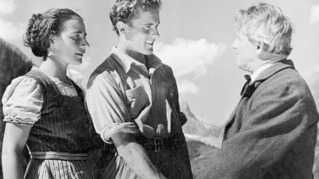 Filmausschnitt. Der Protagonist mit seiner Frau im Hintergrund schüttelt die Hand eines älteren Mannes. Filmausschnitt. Der Protagonist mit seiner Frau im Hintergrund schüttelt die Hand eines älteren Mannes.
