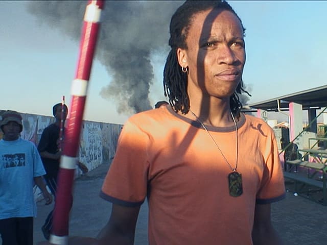 Ein Mann mit orangefarbenem T-Shirt steht auf der Strasse, im Hintergrund steigt grauer Rauch gegen den Himmel. Ein Mann mit orangefarbenem T-Shirt steht auf der Strasse, im Hintergrund steigt grauer Rauch gegen den Himmel.
