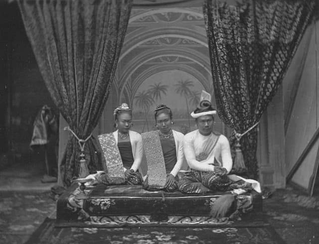Foto von zwei Frauen und dem burmesischen König auf einer Erhöhung, hinten Vorhänge. Foto von zwei Frauen und dem burmesischen König auf einer Erhöhung, hinten Vorhänge.