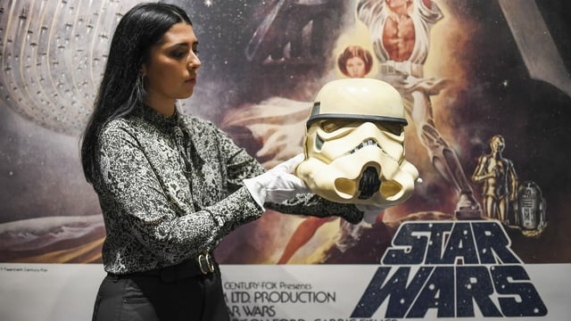 Eine Frau mit Handschuhen hält einen weissen Helm. Im Hintergrund hängt ein Star-Wars-Plakat. Eine Frau mit Handschuhen hält einen weissen Helm. Im Hintergrund hängt ein Star-Wars-Plakat.