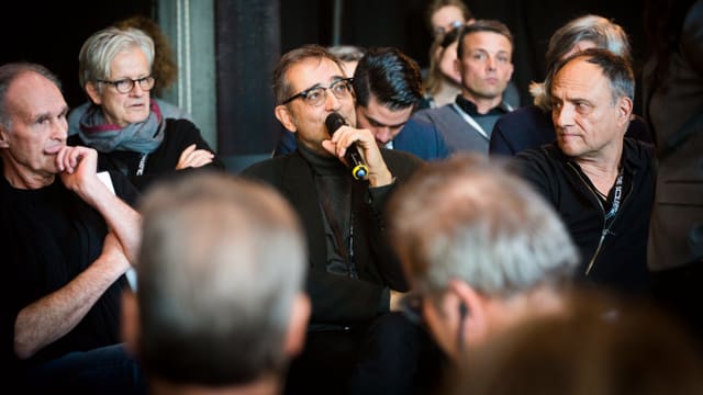 Leute sitzen auf Stühlen in einem Saal. Es wird diskutiert. Leute sitzen auf Stühlen in einem Saal. Es wird diskutiert.