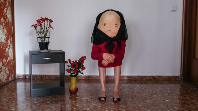 Eine Person mit vornübergebeugtem Rücken, auf dem ein Gesicht gemalt ist, steht in einem kirchenähnlichen Innenraum. Eine Person mit vornübergebeugtem Rücken, auf dem ein Gesicht gemalt ist, steht in einem kirchenähnlichen Innenraum.