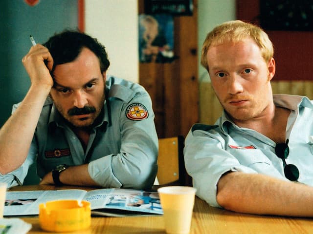 Filmszene: Brenner und Berti sitzen an einem Tisch, sie tragen die Uniformen von Krankenwagenfahrern. Filmszene: Brenner und Berti sitzen an einem Tisch, sie tragen die Uniformen von Krankenwagenfahrern.