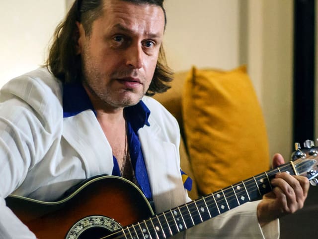Ein Mann mit kinnlangem Haar sitzt auf einem Sofa und spielt Gitarre. Ein Mann mit kinnlangem Haar sitzt auf einem Sofa und spielt Gitarre.