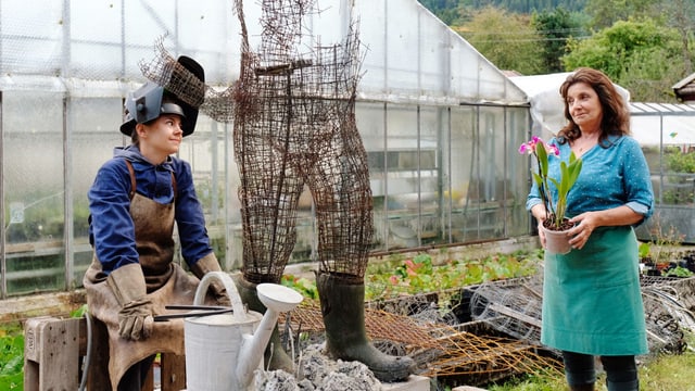 In einem Garten, vor einem Gewächshaus: Eine ältere Frau hält einen Topf mit einer Orchidee in der Hand und schaut eine jüngere Frau an. Diese sitzt neben einer halbfertigen Skulptur einer Person aus Draht. In einem Garten, vor einem Gewächshaus: Eine ältere Frau hält einen Topf mit einer Orchidee in der Hand und schaut eine jüngere Frau an. Diese sitzt neben einer halbfertigen Skulptur einer Person aus Draht.
