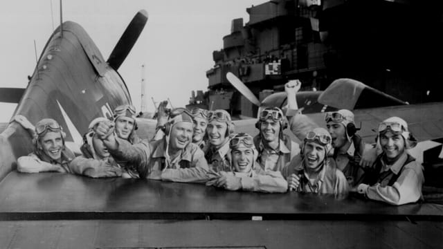 Schwarz-weiss-Aufnahme mit Piloten hinter einem Flügel eines Jagdflugzeugs auf der "USS Lexington", die ihre Abschüsse japanischer Flieger bejubeln. Schwarz-weiss-Aufnahme mit Piloten hinter einem Flügel eines Jagdflugzeugs auf der "USS Lexington", die ihre Abschüsse japanischer Flieger bejubeln.