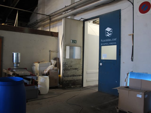 Eingangstüre mit dem Logo von FluidSolids, Grosse Plastikkübel, Gerätschaften und Kartonschachteln stehen herum. Eingangstüre mit dem Logo von FluidSolids, Grosse Plastikkübel, Gerätschaften und Kartonschachteln stehen herum.
