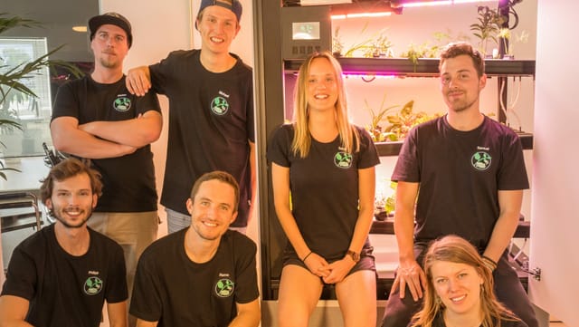 Ein Gruppe von sieben Personen steht und kniet vor einem schwarzen Gerät, in dem Pflanzen wachsen. Ein Gruppe von sieben Personen steht und kniet vor einem schwarzen Gerät, in dem Pflanzen wachsen.