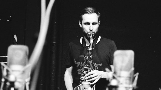 Ein Mann in T-Shirt spielt Saxophon vor zwei Mikrofonen (s/w-Bild).