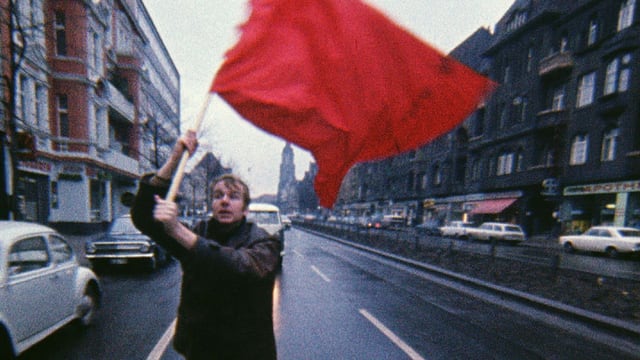 Ein Mann trägt einer rote Fahne. Er rennt auf einer Strasse.