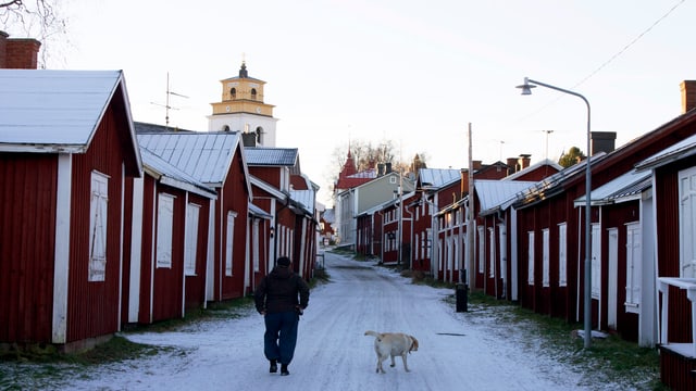 Mann mit Hund auf einem verschneiten Weg, links und rechts rot gestrichene Holzhäuser. Mann mit Hund auf einem verschneiten Weg, links und rechts rot gestrichene Holzhäuser.