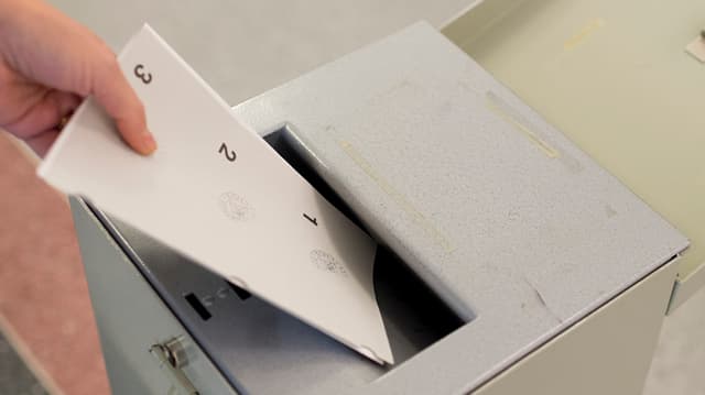 Eine Person wirft Unterlagen in einer Wahlurne Eine Person wirft Unterlagen in einer Wahlurne