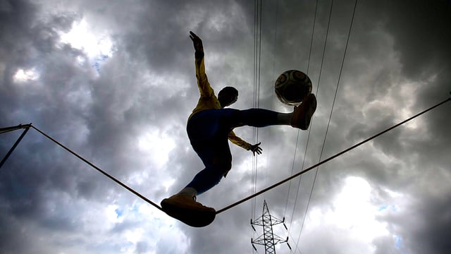 Junge balanciert mit einem Ball auf einem Seiltanz-Seil. Im Hintergrund ist ein düsterer Wolkenhimmel zu sehen.