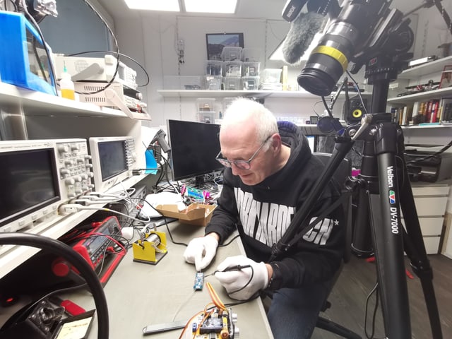 Andreas Spiess sitzt an seinem langen Labortisch und lötet an einem Elektronikbauteil herum. Rechts eine Kamera, die alles filmt. Andreas Spiess sitzt an seinem langen Labortisch und lötet an einem Elektronikbauteil herum. Rechts eine Kamera, die alles filmt.