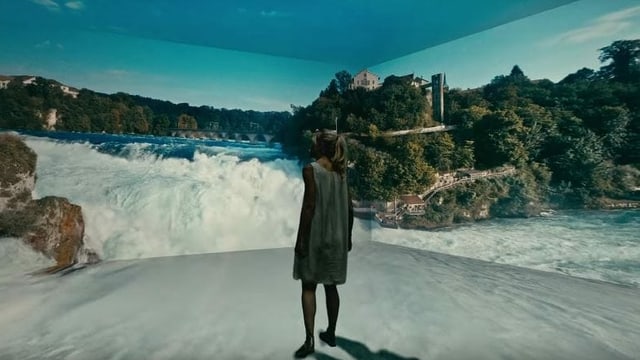 Eine Frau steht in einem Raum, auf dessen Wand der Rheinfall projiziert wird. Eine Frau steht in einem Raum, auf dessen Wand der Rheinfall projiziert wird.