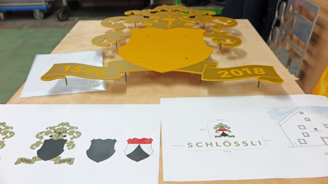 Metallwappen für Schlössli Wohlen auf einem Tisch, davor Planzeichnung für Installation des Wappens Metallwappen für Schlössli Wohlen auf einem Tisch, davor Planzeichnung für Installation des Wappens