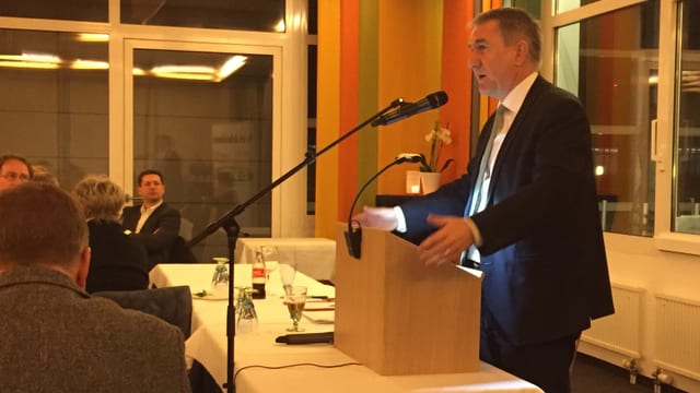 Mann in Anzug an Rednerpult, davor Köpfe. Mann in Anzug an Rednerpult, davor Köpfe.