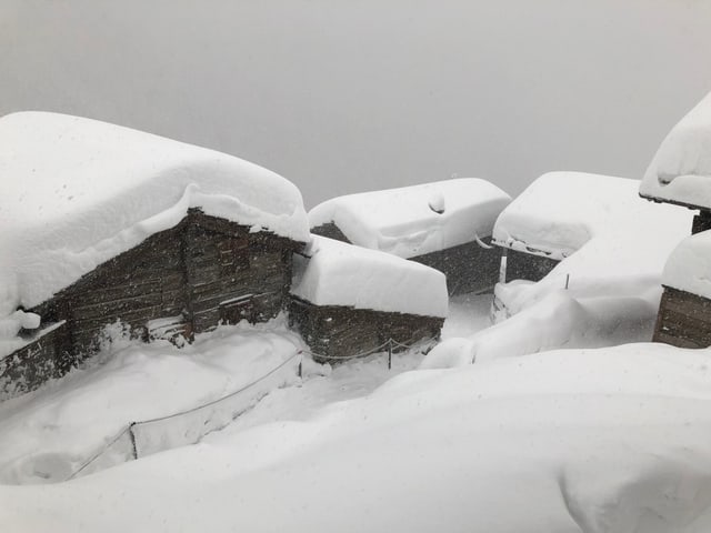 Bergdorf mit 5 Häusern versinkt im Schnee. Es hat meterhoch Neuschnee. Der Himmel ist grau und alles weiss.