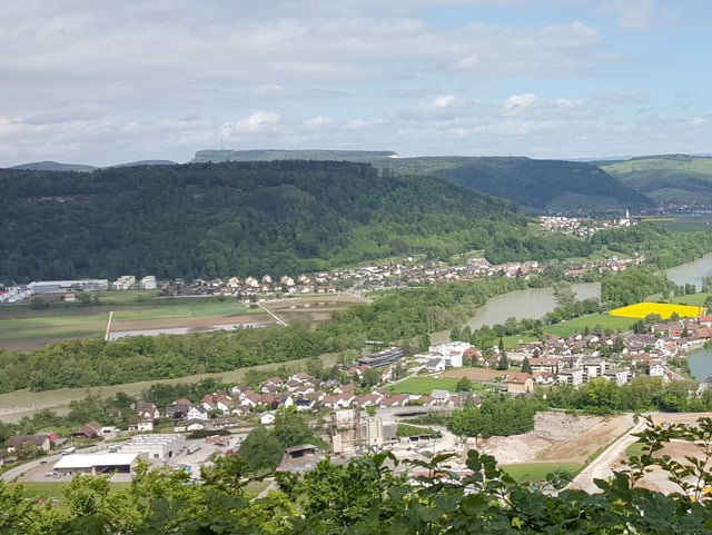 Am Himmel teilen sich kleine Quellwolken und blaue Flecken den Platz am Himmel. Die Landschaft ist geprägt von bewaldeten Hügeln. In der Mitte des Bildes ist zwischen Bäumen die braune Aare zu erkennen. Dahinter ein Acker mit Wasserlachen darauf.