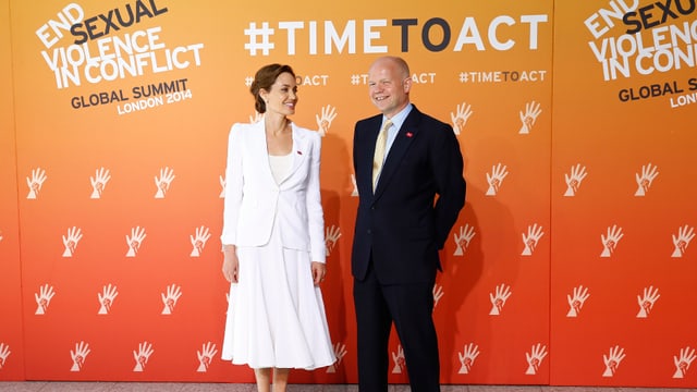 Jolie in Weiss und Hague im Anzug posieren vor einer Wand, auf der steht «end sexual violence in conflict»