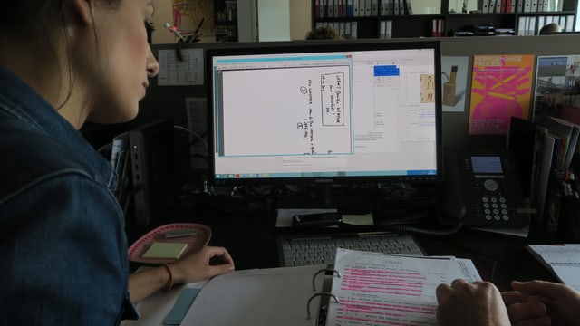 Eine Frau links im Bild schaut auf einen Ordner, der auf einem Pult liegt. Hinter dem Ordner steht ein Computerbildschirm. Eine Frau links im Bild schaut auf einen Ordner, der auf einem Pult liegt. Hinter dem Ordner steht ein Computerbildschirm.