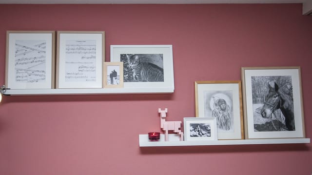 Das Bild zeigt die rosarote Wand mit zwei Bildleisten. Die verschiedenen Bilder darin sind unterschiedlich gross und in weissen und hölzernen Rahmen. Das Bild zeigt die rosarote Wand mit zwei Bildleisten. Die verschiedenen Bilder darin sind unterschiedlich gross und in weissen und hölzernen Rahmen.