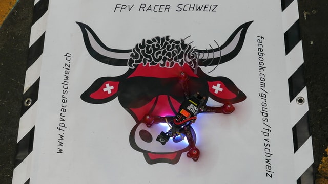 Das Logo der FPV-Racer Schweiz (roter Stierkopf mit Schweizerkreuz in den Ohren und coller Sonnenbrille), darauf eine Racer-Drohne. Das Logo der FPV-Racer Schweiz (roter Stierkopf mit Schweizerkreuz in den Ohren und coller Sonnenbrille), darauf eine Racer-Drohne.