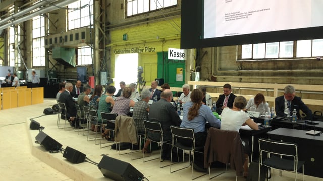 Menschen sitzen in einer Halle an einem langen Tisch, auf einer Leinwand wird das Diskussionsthema gezeigt. Menschen sitzen in einer Halle an einem langen Tisch, auf einer Leinwand wird das Diskussionsthema gezeigt.